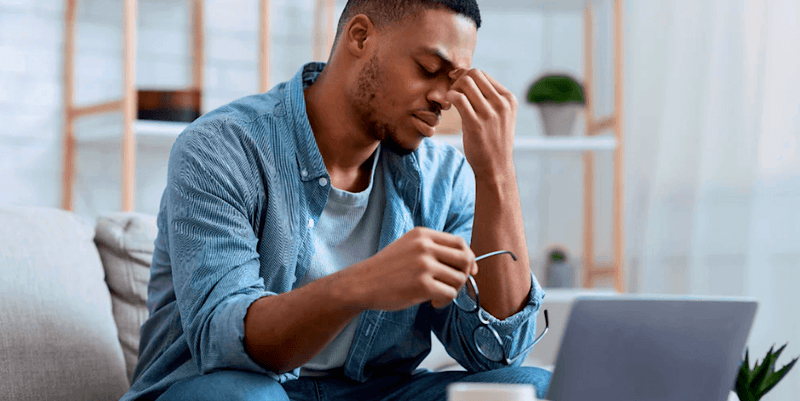 A man at a laptop taking off his glasses and pinching the bridge of his nose.