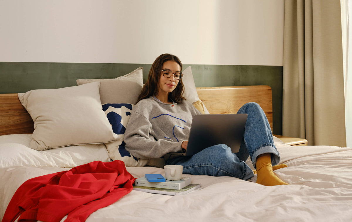 A woman sitting on a bed working on a laptop, wearing glasses.