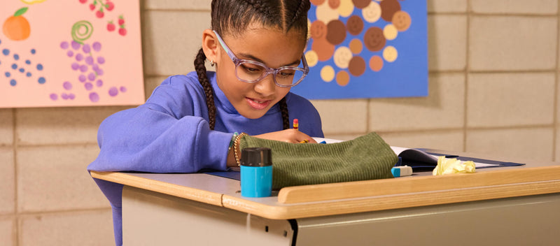 A young girl wearing glasses drawing at a classroom desk.
