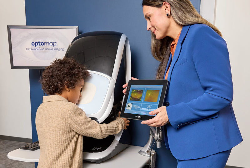 A young child looking into a retinal imaging scanner while an optometrist shows the results on a tablet.